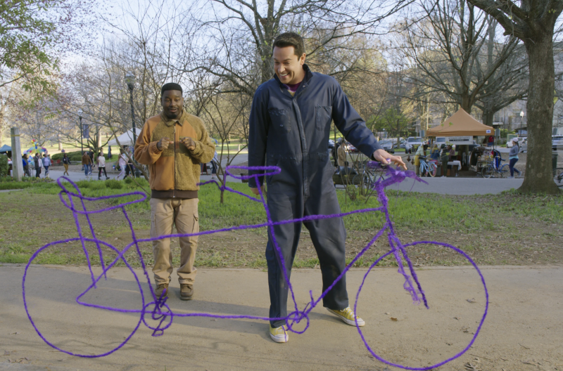 A white man in a boiler suit holds the handlebars of a life-size stick drawing of a bicycle. A Black man looks on looking surprised and delighted.